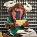 Portrait of the New College mascot Goliath Gnu (a wildebeest dressed in a green New College jersey) sitting and reading a letter, with a large bag of letters by his feet and a wall of mail slots behind him.