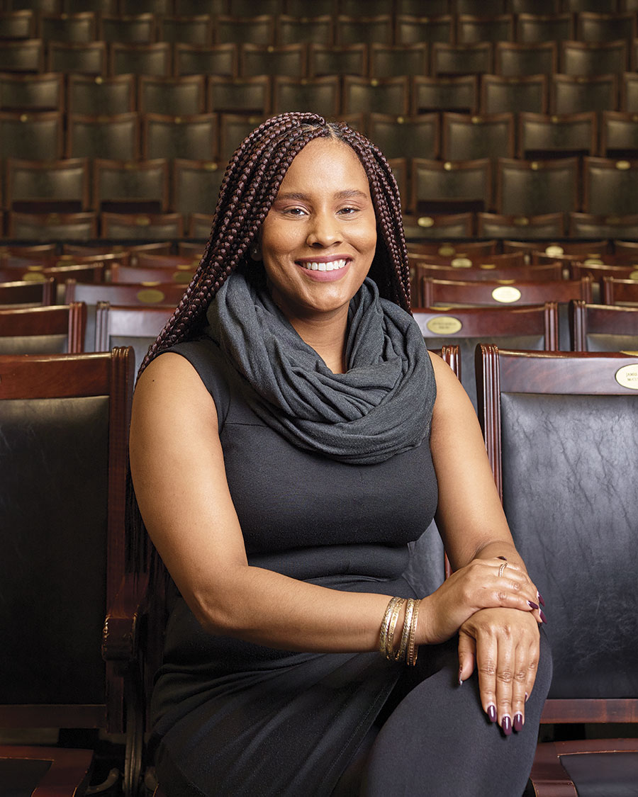 Portrait of a seated, smiling woman with box braids and folded hands [Lydia Gill in U of T’s Convocation Hall]; rows of seats visible behind her.