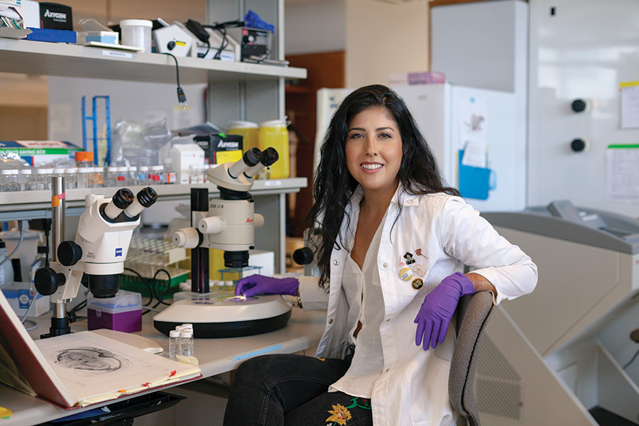 Portrait of a scientist with long dark hair [Samantha Yammine] in a lab coat next to microscopes and an open brain atlas.